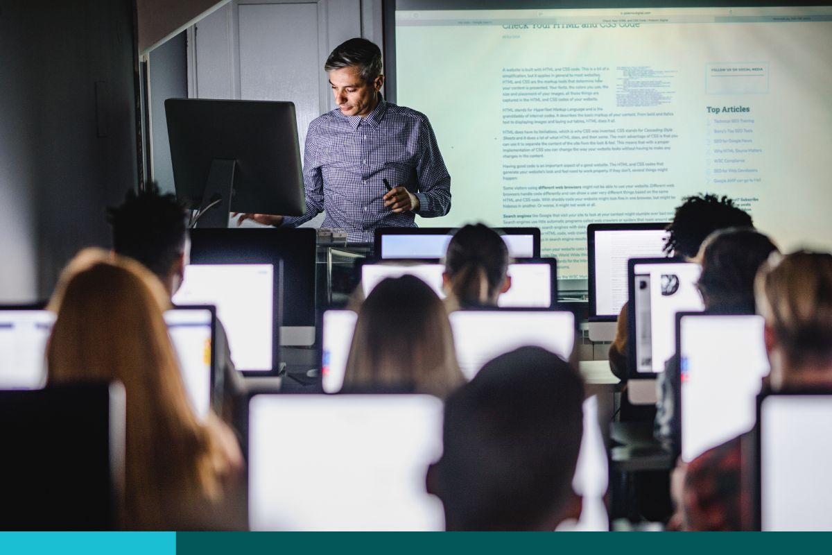 Teacher presenting in front of his class. All students have screens. On projector screen is content about HTML and CSS